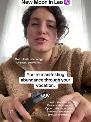 A woman with curly dark hair in a brown fuzzy jacket is speaking directly to the camera. Text overlays include a title about the new moon in Leo along with motivational phrases and affirmations. She is holding a deck of cards.