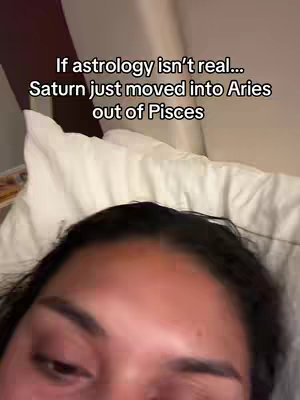 A selfie-style shot of a person lying in bed, viewed from the chest up. The person's face is close to the camera, with the upper half of the face visible, with dark hair and an expression of mild surprise. Behind them is a white pillow and a hint of a wall. Text overlayed on the image reads, 'If astrology isn't real... Saturn just moved into Aries out of Pisces'.