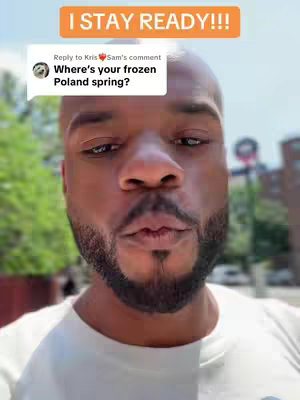 A close-up shot of a Black man with a shaved head and a well-groomed beard. He's looking towards the camera with a neutral expression, his lips slightly pursed as if he's about to speak or has just tasted something. In the background, a blurred city scene is visible, with green foliage and building structures suggesting an outdoor setting. White text box overlays with some texts above the man's head.