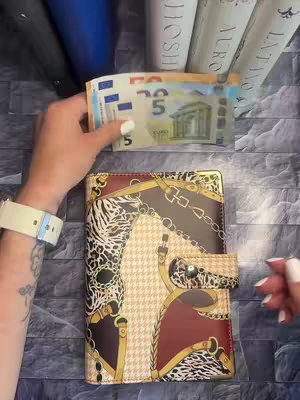 A person's hands are holding a wallet and a Euro banknote. The wallet is a patterned accessory with a clasp and other embellishments. Euro banknote is held partially next to the wallet.