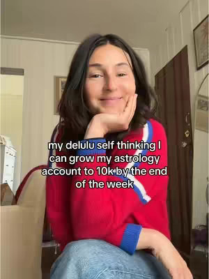 A young woman with dark hair, is sitting in a room holding her hand near her face and smiling. She's wearing a red sweater with blue and white stripes. The background includes a light-colored wall, a door, and framed pictures.