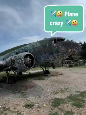 An aging, graffiti-covered airplane is the central focus, parked on a gravelly field under a partly cloudy sky. A light green speech bubble overlay features the text 'Plane crazy' with airplane and smiling face emojis.