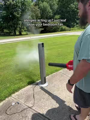 A man is outside on a sunny day, pointing a leaf blower at a silver fan. The fan is standing on a concrete surface next to a patch of grass. The leaf blower is red and black and is blowing a cloud of dust towards the viewer. The man is wearing a light green t-shirt, dark shorts, and sandals. In the background, there are trees and a road.