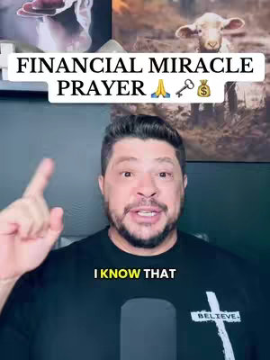 A man with a beard and short dark hair is pointing upwards with his right index finger against a wall. Above him is text in a white box saying FINANCIAL MIRACLE PRAYER and some emojis. He is wearing a black Believe t-shirt and the background is dark.