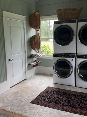 An interior shot of a laundry room or utility area shows a door on the left and white appliances and shelving on the right, with a patterned rug in the foreground. The walls are light green vertical paneling. The floor has a light-colored, grey, herringbone pattern. The appliances, including a set of stacked washers and dryers, are white with black control panels and are positioned next to a window that shows an out-of-focus green yard. Two large wicker baskets are mounted on the walls between the door and the window. A smaller wicker basket sits atop the stacked machines.