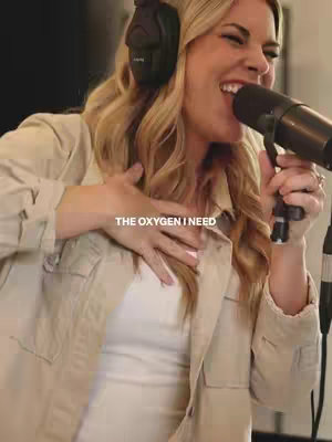 A woman with blonde hair stands in a recording studio, singing into a microphone. She has a headset on and is wearing a light jacket over a white top. The text overlay reads, 'THE OXYGEN I NEED.'