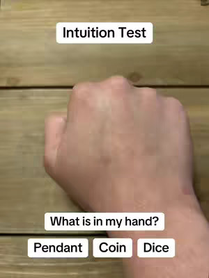 A close-up on a wooden surface with a hand in the center. The hand is closed, and the image is overlaid with text boxes at the top and bottom. The top text says 'Intuition Test,' and the bottom has the words 'Pendant,' 'Coin,' and 'Dice' as options.