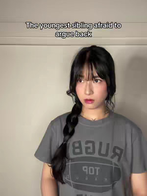A young woman with dark hair in a braid and bangs looks off to the side with a slight grimace, wearing a grey t-shirt. The background is a plain, light-colored wall.