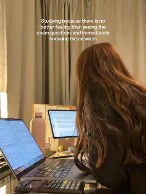 A woman with long brown hair sits at a desk with two laptops and a water bottle, studying. Text overlayed on the image reads: Studying because there is no better feeling than seeing the exam questions and immediately knowing the answers.