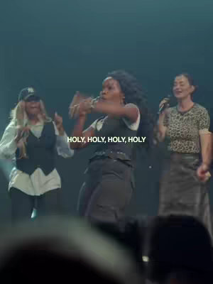 The cover image shows three women on a stage with a dark background. The center woman is the main focus and is singing while gesturing with her hands. The other two women, one on each side, are also singing, with microphones at hand. The background appears to be a stage setting with dim lighting. A text overlay displays the words "HOLY, HOLY, HOLY, HOLY".