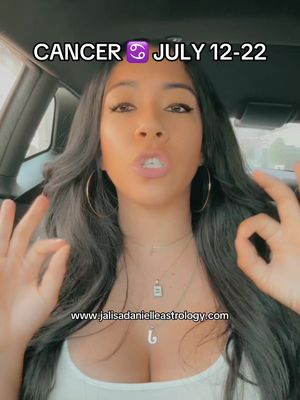 A woman is seated in a car's driver's seat, facing forward and speaking. Above her, text reads "CANCER ♋ JULY 12-22". The background appears to be out of focus, suggesting an interior car setting. She is wearing a white top and multiple necklaces, with large hoop earrings adding to her appearance. She's using hand gestures. Low in the frame, is website text.