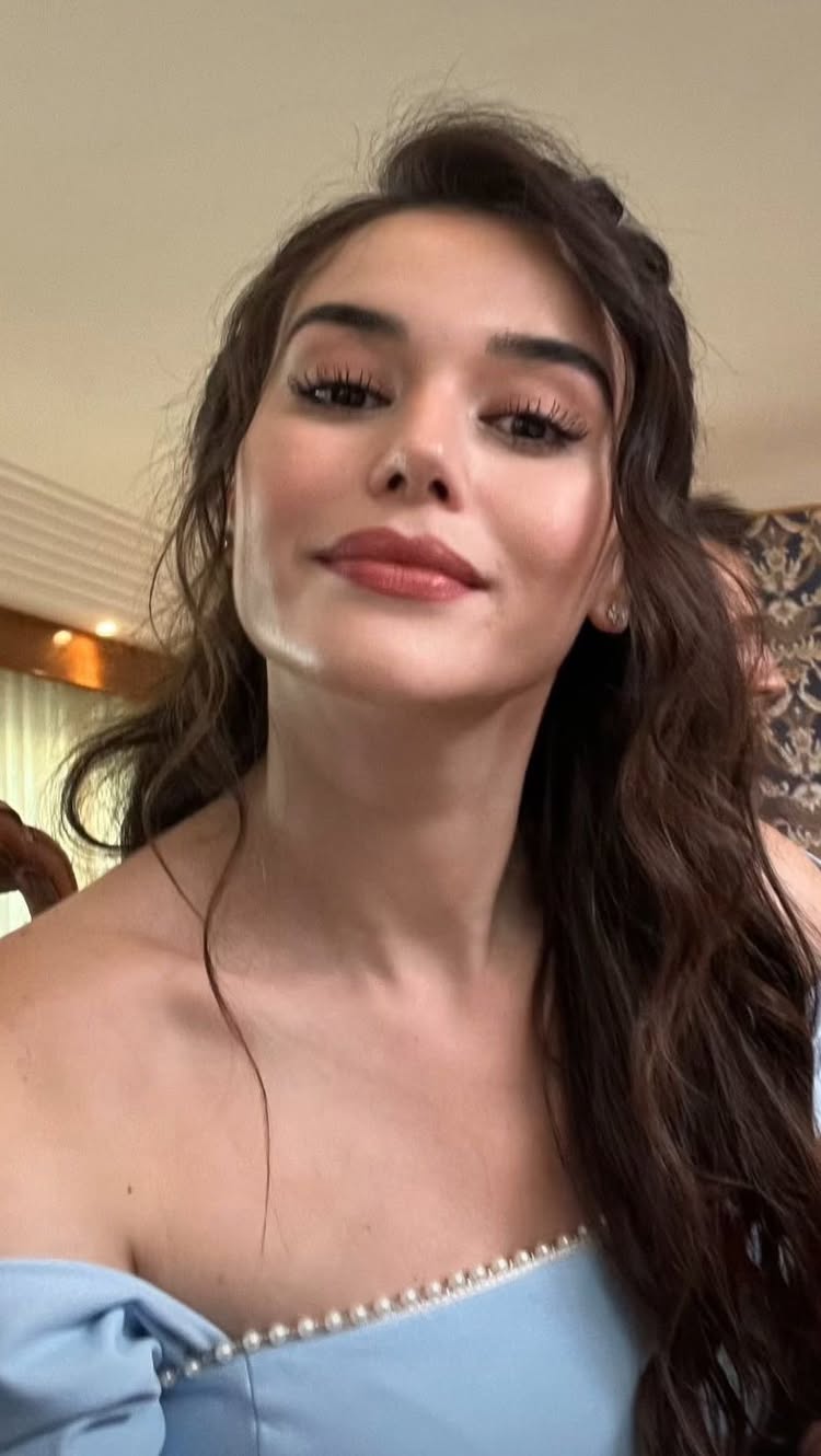 A selfie-style, vertical shot features a fair-skinned woman with long, wavy brown hair. She is wearing a light blue off-the-shoulder top with a pearl trim. The woman's gaze is directed towards the camera, and she has soft, natural-looking makeup with a nude lipstick shade. The background is slightly blurred, featuring a warm-toned, ornamental wall.