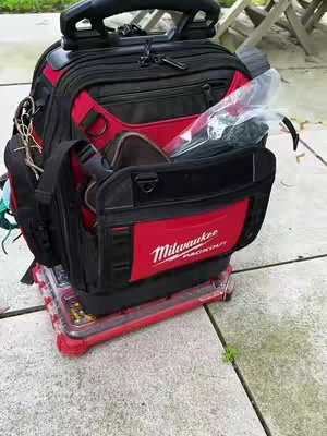An overhead shot of a red and black tool bag on a red rolling base, set on a grey stone surface. The bag has the 'Milwaukee PACKOUT' logo prominently displayed. The background shows a glimpse of green plants and a wooden structure.