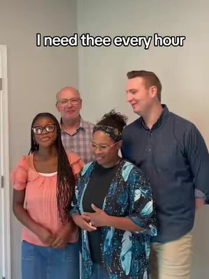 A group of four people indoors posing for a photo. The individuals consist of two men and two women, standing close together. The background is a neutral light gray wall. The text overlay on the top says, "I need thee every hour."