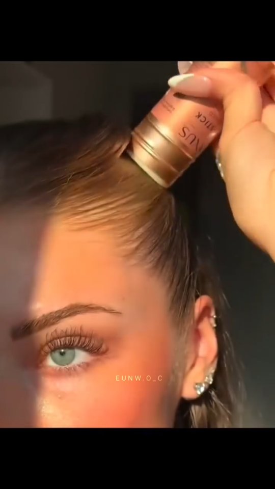 Close-up shot of a woman partially shown with a makeup product in use. The right side of her face is in view, with an eye visible reflecting sunlight. The hair is styled away from the face, and part of a makeup applicator is being used near the hairline.