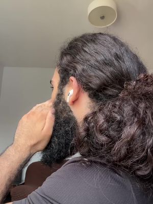 A man with a long, dark beard and curly hair is seen from the side, wearing an earpiece. He is holding his hand up to his face in a gesture of surprise, near a light-colored wall and a circular ceiling light. The image is well-lit naturally.