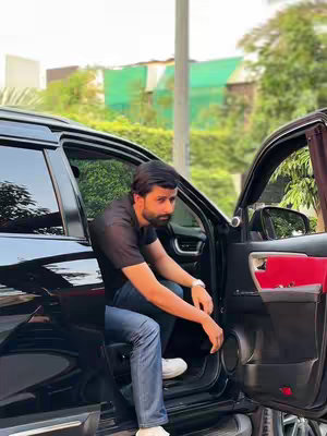 A man in a black t-shirt and blue jeans is seated inside a black car with the door open. The car is parked outdoors with green trees and foliage in the background.