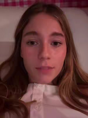 A close-up shot of a young woman's face, centered in the frame. She has long, light brown hair and a fair complexion. She wears a white collared shirt, with a few buttons done up. The background is a soft, blurry pattern of pink and white checks, suggesting a bedroom or similar setting.