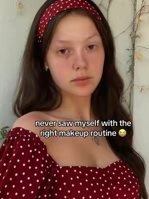 A young woman with long, dark hair and a maroon polka-dot headband is the focus of the image. She is gazing towards the camera with a somewhat neutral expression. Her skin appears to be fair. She is wearing a red top with white polka dots, revealing her shoulders. The background consists of a plant and an off-white wall.