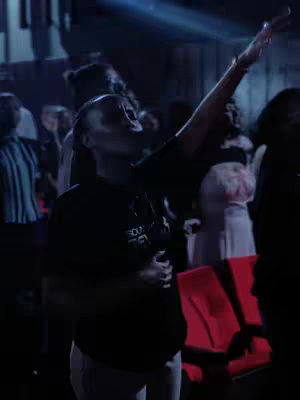 A woman with dark skin and short hair is in a crowded space with her arm and hand raised, seemingly in worship. Others are visible in the background, suggesting a religious or performance setting. The lighting is dim, with a spotlight or similar source illuminating portions of the scene.
