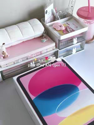 An overhead shot of a desk setup with a pink iPad box and various accessories. The composition is aesthetically pleasing with a focus on pink and white tones. The iPad box is open, revealing the device. Other elements include desk organizers, a pink tumbler, and decorative items, suggesting a study or creative space.