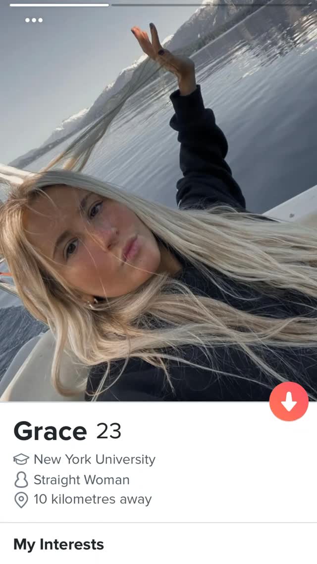 A selfie-style shot of a young woman with long blonde hair on a boat with a scenic background of water and mountains. She's holding up two fingers in a peace sign, smiling with her head slightly tilted. In the lower portion, a dating app profile is visible with basic information.