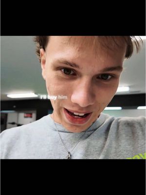 A mid-shot of a young man with light skin looking directly at the camera and speaking. Text on the image reads, 'I'll buy him.' The man wears a light grey t-shirt and a silver chain necklace. The background is a slightly blurred interior space, likely a room.