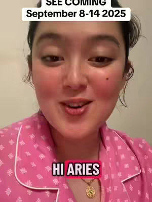 A woman with a light complexion and dark hair is centered in a close-up shot. She is wearing a pink, patterned pajama top with a white collar. Text overlays include "SEE COMING September 8-14 2025" at the top and "HI ARIES" in a pink box at the bottom. The background is a neutral, light-colored wall. She has a warm, slightly rosy complexion.