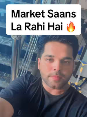 A selfie-style video cover image featuring a man in a black t-shirt, against a domestic setting. The man, with dark hair and a focused expression, is positioned in the foreground. Above him is a white box with the overlaid text in Hindi, the text is in bold, set against a blurred background composed of possibly stairs and a scooter.
