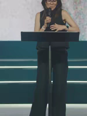 A woman with dark hair, glasses and a black outfit is standing behind a podium. She is holding a microphone and appears to be speaking. The background has a blurred abstract pattern.
