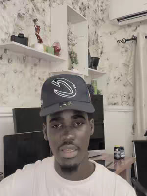A young Black man wearing a cap and a white t-shirt, is positioned in front of a wall with a distinctive marbled pattern. Shelves and an air conditioning unit are visible in the background. The overall lighting is bright, and the composition is a medium shot focusing on the upper body of the man.
