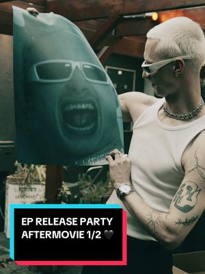 The cover image presents a man holding a poster featuring a distorted, graphic close-up of a face with sunglasses and an open mouth. The man is fair-skinned with short, bleached blonde hair and sunglasses. He wears a white tank top and a chain necklace, and has a visible tattoo on his arm. The background is blurred, suggesting an outdoor setting. Overlayed text at the bottom reads "EP RELEASE PARTY AFTERMOVIE 1/2 🖤", highlighting the event and serving as a title.