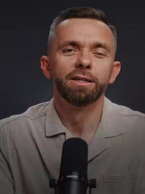 A medium shot features a man with short hair, a trimmed beard, and a light-colored collared shirt. He looks directly at the camera, with a slightly open mouth as if speaking. A black microphone is positioned in front of him, and the background is out of focus, creating a studio-like setting.
