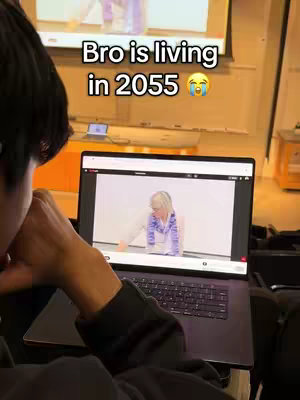 A person is sitting in a dark room with a hand in the frame, looking at a laptop. The laptop screen shows an older woman presenting a class. The background has an orange object and an image projected on a screen. The text overlay states: "Bro is living in 2055 😭"