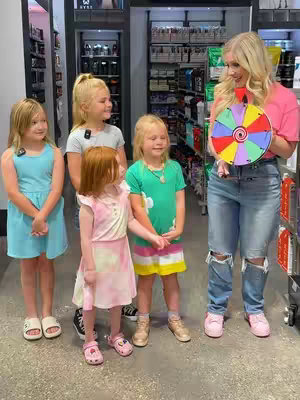 A group of five girls, all with blonde hair, are standing in a store setting. A woman, holding a colorful spinning wheel, stands at the right edge, smiling and looking at the girls. There are shelves of products visible.