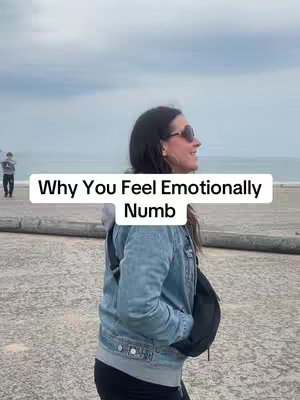 A woman stands sideways on a beach, looking towards the right, with a cloudy sky in the background. She wears sunglasses, a denim jacket, and a black bag across her body. A text overlay reads 'Why You Feel Emotionally Numb'. In the background, a person is casually walking.