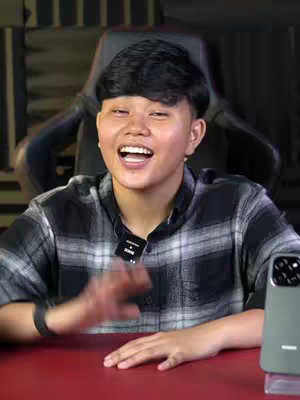A young woman with short, dark hair and a plaid shirt smiles and gestures while seated in front of a black and red background. A phone is visible on the right side of the frame.
