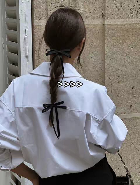 A woman with long brown hair is seen from the back, wearing a white collared shirt and dark pants. Her hair is styled into a braid, adorned with black bows at the top and bottom of the braid. There is also a black bag hanging by a chain in the image. The overall composition is simple and focuses on the woman's back and hair detail. The background is blurred, showing a wall of light color.