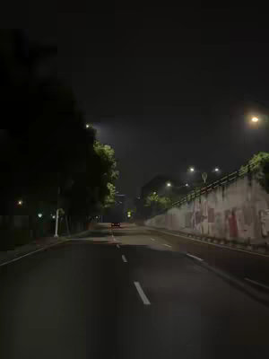 A night scene captures a road disappearing into the distance under an overcast sky. Streetlights line the route, and trees are visible on the left. In the center, a car's taillights hint at movement, and a concrete barrier with graffiti runs along the right side of the road.