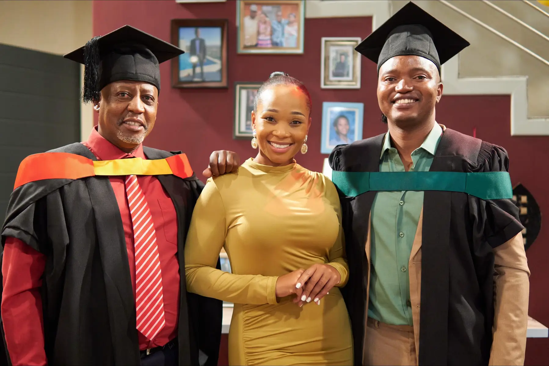 The image features three people in a room. Two men and one woman standing closely together. All three are smiling at the camera and wearing graduation attire. The woman is centrally positioned with her hands clasped in front of her. Behind them, there are framed pictures on a burgundy wall.