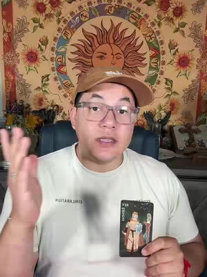 A man with glasses and a brown cap is holding a tarot card while speaking to the camera. The background features a tapestry with a sun design and floral patterns.
