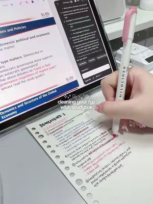 The image shows a person writing notes on a piece of paper with a pink and white highlighter, with a tablet displaying study materials in the background. The scene is set on a desk with other school supplies.