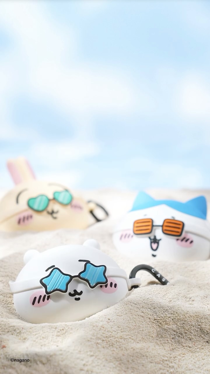 The image shows three cartoon earbuds cases resting in sand, with a blue sky backdrop. The cases are designed to resemble characters: a white bear-like figure with star-shaped sunglasses, a cat with blue ears and orange-lensed sunglasses, and a rabbit with heart-shaped sunglasses. The cases are in focus and the sand and background sky are slightly blurred.