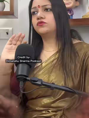 A woman with long dark hair and fair skin is shown in a medium shot, positioned slightly off-center, facing towards the left with an upward gaze. She is wearing a gold-colored sari with a red bindi on her forehead. A microphone is positioned in front of her, suggesting a recording. The background includes a framed picture and a figurine, hinting at an indoor setting, possibly a podcast studio. Text across the lower left reads, "Credit by - Himanshu Sharma Podcast".
