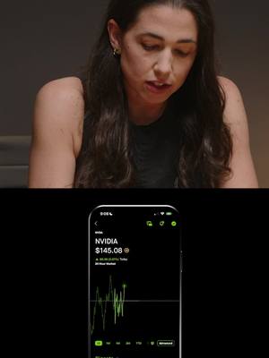 The image features a woman looking down, with the background of the scene partly visible, in front of a smartphone screen displaying a stock chart. The phone screen is black with green highlights, showing financial data and the stock price of "NVIDIA". The woman has long dark hair and is wearing a sleeveless top, with a neutral expression. The background is a dark, blurred setting.