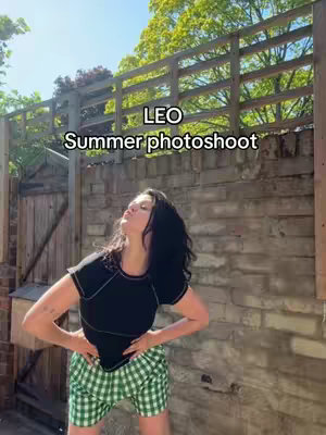 A woman poses outdoors with a brick wall and fence behind her. She is wearing a black top, green checkered shorts, and her hair is down. The text overlay says 'LEO Summer photoshoot'.