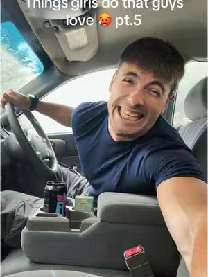 A man with short brown hair wearing a navy blue t-shirt is sitting in the driver's seat of a car in a selfie-style shot. He is smiling widely, with his teeth visible, and holding the steering wheel. The interior of the car is visible in shades of gray and beige.