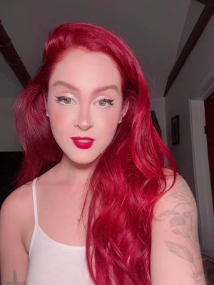 A selfie-style shot of a woman with vibrant red hair, styled in a voluminous, wavy fashion. She has fair skin, and soft makeup including prominent red lip and white eyeliner. She wears a white spaghetti-strap top. The background is softly lit, revealing parts of the interior of a room, including a door frame and architectural details.