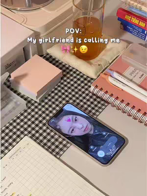 A top-down shot featuring a table surface with a black and white checkered pattern. On it are various items: a smart phone displaying a selfie of a person, a glass of amber liquid, a stack of pink sticky notes, a pink notebook, a pen, and a pad of paper. The phone shows the selfie with a pink heart graphic over the subject's forehead. Overlayed on the scene is the text, 'POV: My girlfriend is calling me' in white text with a pink bow and a smiling emoji.