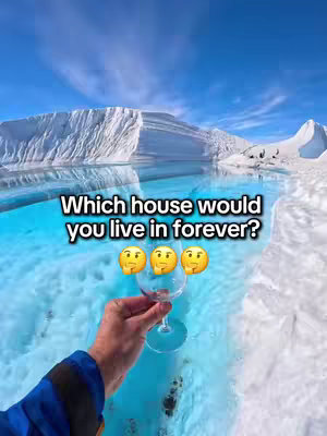 The image shows a hand holding a wine glass against a backdrop of glacial ice and a clear blue sky. The hand, wearing a blue sleeve, is positioned in the foreground, with the wine glass as the central element. Above the glass are three emojis, and the background features icy formations and clear water. Text overlay reads: 'Which house would you live in forever?'
